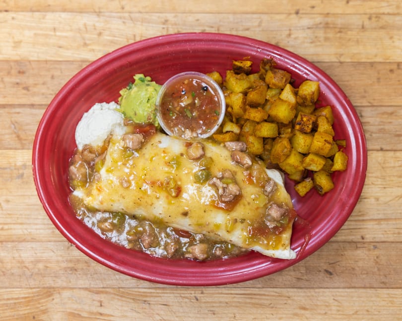 A breakfast burrito smothered in green chili sauce, with a side of crispy potatoes, guacamole, and sour cream.