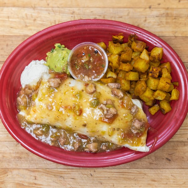 A breakfast burrito smothered in green chile sauce, with a side of potatoes, guacamole, sour cream, and salsa.