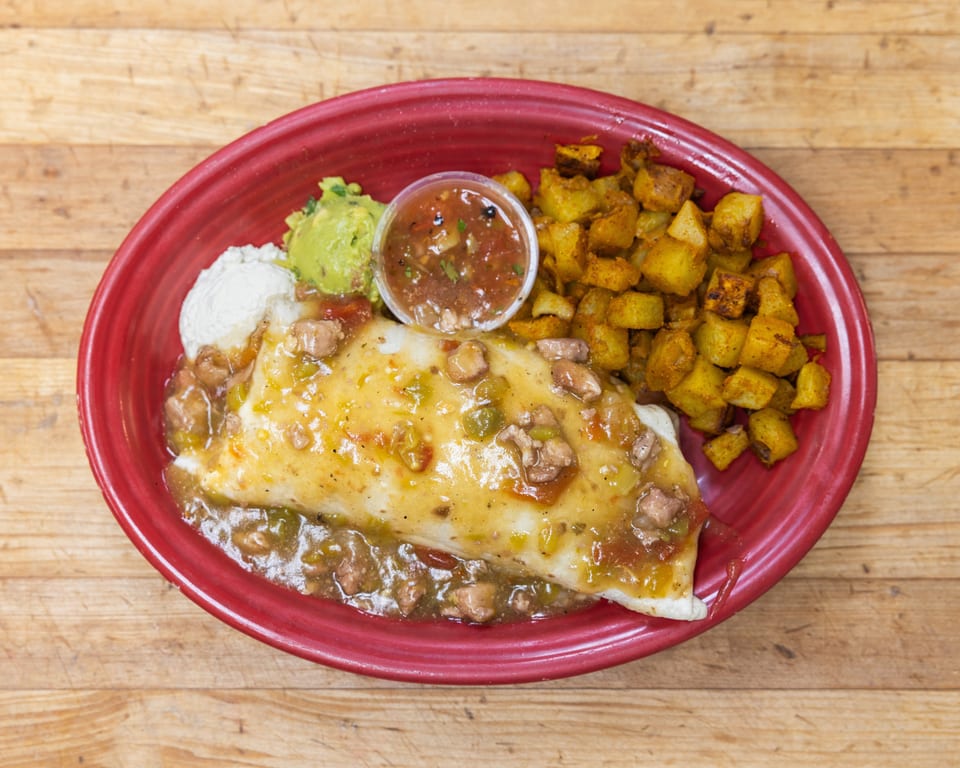 A breakfast burrito smothered in green chile sauce, with a side of potatoes, guacamole, sour cream, and salsa.