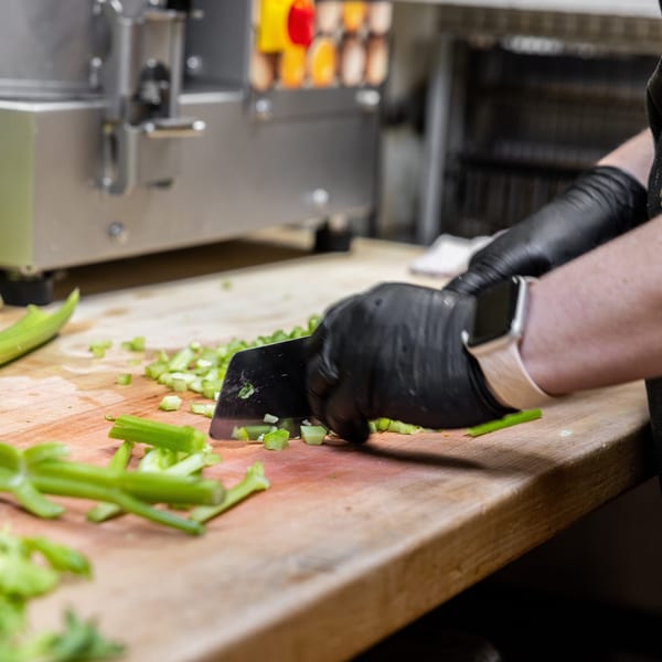 Chef in black gloves chopping celery on a wooden cutting board in a commercial kitchen.