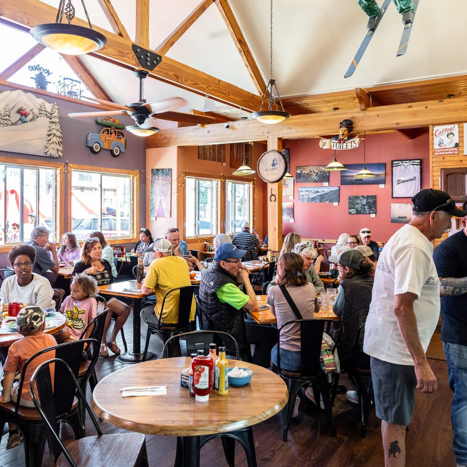 A bustling restaurant interior with many patrons seated at tables, decorated with mountain-themed art and skis.