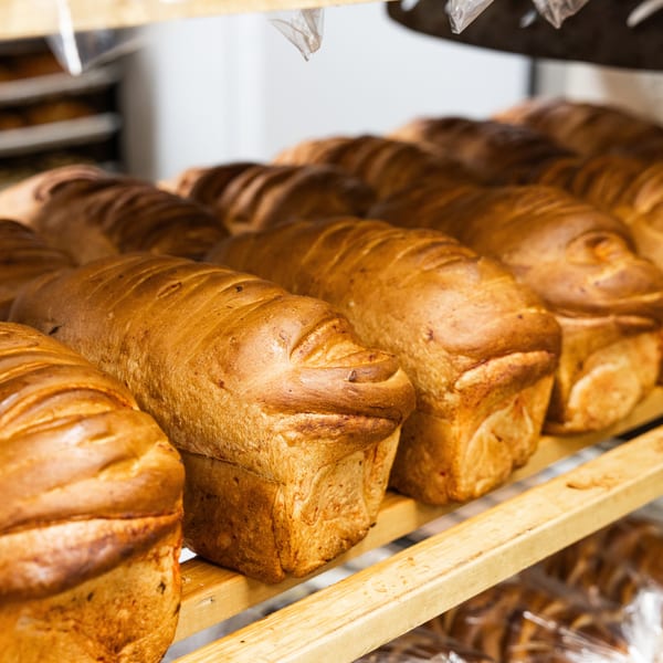 Rows of freshly baked, golden-brown bread loaves sit on wooden shelves in a bakery.