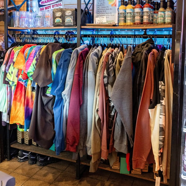 A rack of colorful tie-dye and solid-colored shirts for sale, with drinks and sauces nearby.