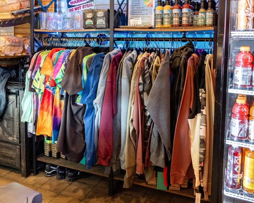 A rack of colorful tie-dye and solid-colored shirts for sale, with drinks and sauces nearby.