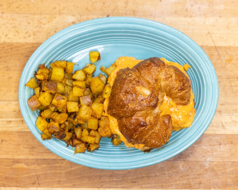 Croissant sandwich with creamy orange filling and a side of crispy roasted potatoes on a blue plate.