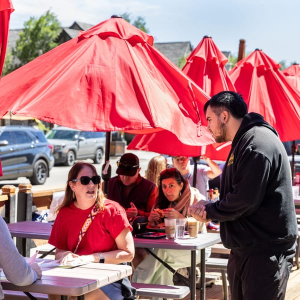 A server talks to customers seated at outdoor tables under red umbrellas on a sunny day.
