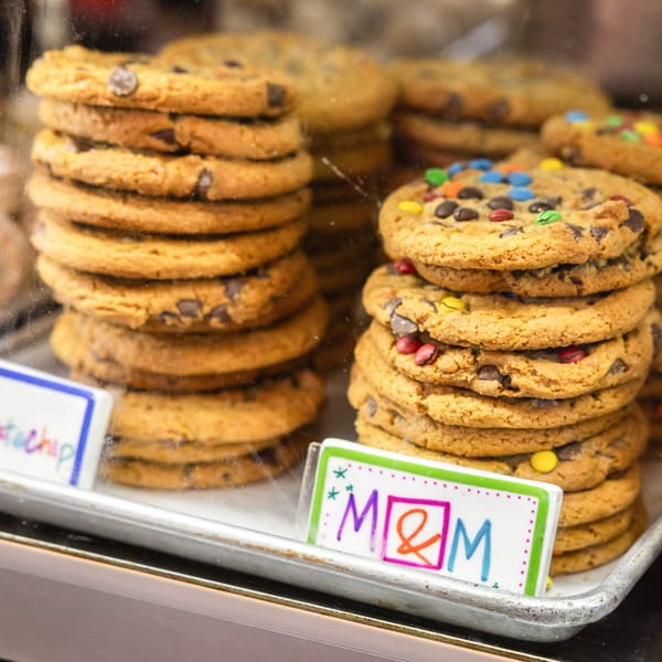 Stacks of chocolate chip and M&M cookies displayed in a glass bakery case.