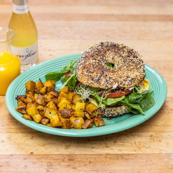 Everything bagel sandwich with spinach, tomato, sprouts, and avocado, served with roasted potatoes and drinks.