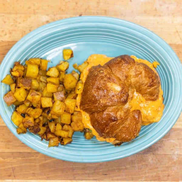 Croissant sandwich with creamy orange filling and a side of crispy breakfast potatoes on a blue plate.